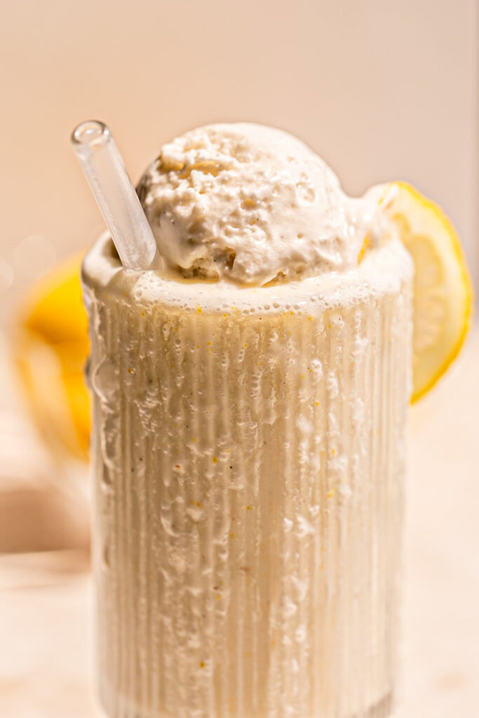 a close up of an overflowing glass of lemonade milkshake with a scoop of vanilla ice cream on top, a glass straw sticking out and a slice of lemon on the glass