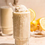 two glasses of lemonade milkshake, one overflowing down the glass topped with a scoop of vanilla ice cream and a bowl of lemons in the background