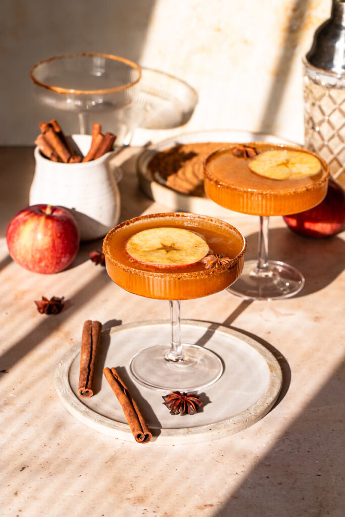 two glasses of pumpkin apple cider mocktail with a jar of cinnamon sticks, two apples, star anise in the background
