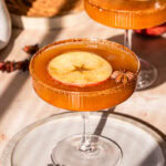 a close up of pumpkin apple cider mocktail with a slice of apple and star anise in the glass, a plate of coconut sugar in the background