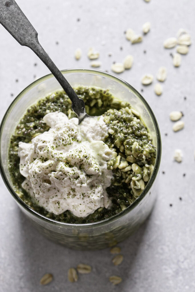 an overhead shot of Oat Milk Matcha Latte Overnight Oats with a spoon in the glass topped with oat milk dairy free whipped cream