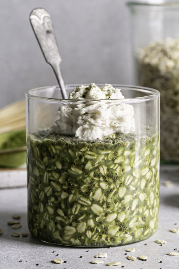 a close up of Oat Milk Matcha Latte Overnight Oats in a glass with a spoon sticking out of it, topped with oat milk whipped cream
