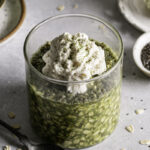 3/4 shot of Oat Milk Matcha Latte Overnight Oats in a glass topped with oat milk whipped cream and a spoon beside the glass