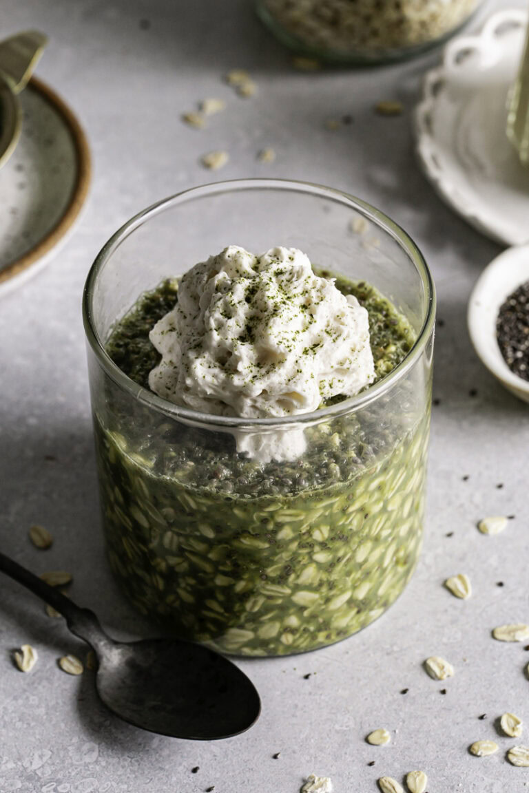 3/4 shot of Oat Milk Matcha Latte Overnight Oats in a glass topped with oat milk whipped cream and a spoon beside the glass