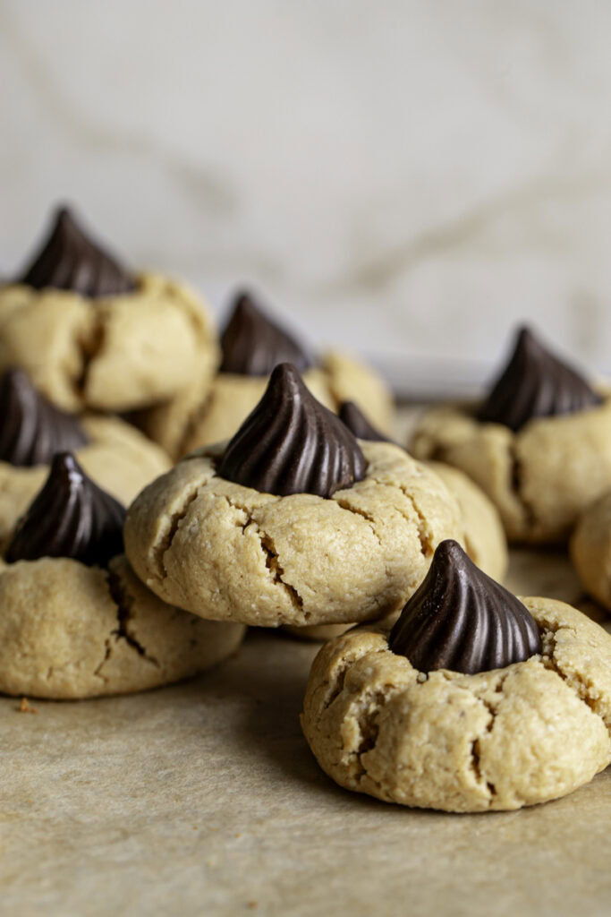 a close up of three Almond Flour Peanut Butter Blossom Cookies stacked on top of one another on a piece of parchment paper, other cookies in the background.