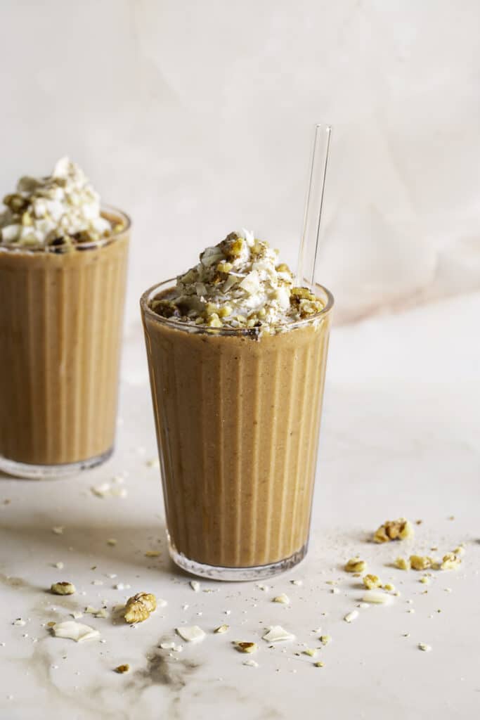 two glasses of carrot cake protein smoothie topped with oat whipped cream garnished with toasted coconut and chopped walnuts.
