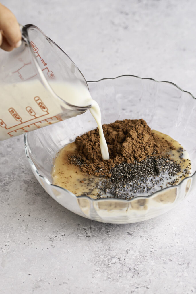 action shot pouring plant milk from a glass measuring cup into the bowl with the chia seeds and cacao powder.