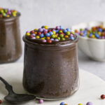 close up shot of a jar of Cosmic Brownie Chocolate Chia Seed Pudding with a bowl of candy coated rainbow chocolate chips and another jar of chia pudding in the background.