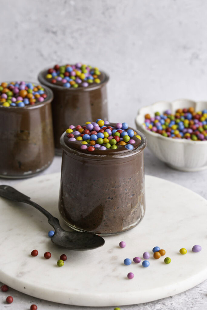 three jars of Cosmic Brownie Chocolate Chia Seed Pudding with a bowl of candy coated rainbow chocolate chips in the background.