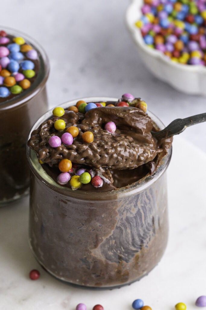 a jar of Cosmic Brownie Chocolate Chia Seed Pudding with a spoon scooping some out of the jar resting on the mouth of the jar.