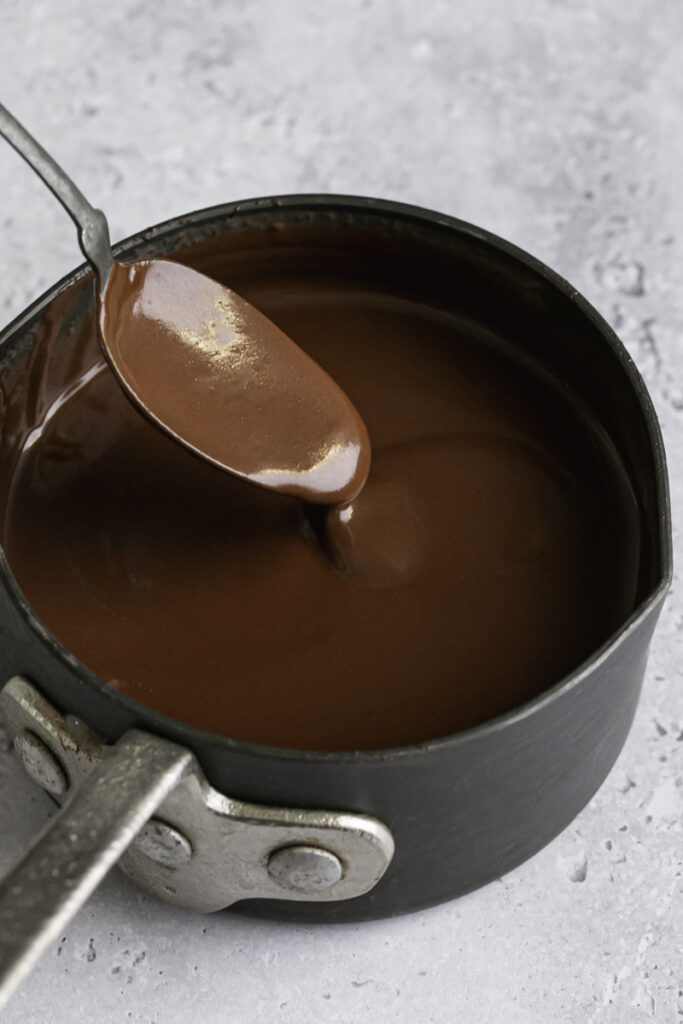 melted smooth and silky chocolate ganache in a small saucepan with some ganache drizzling off a spoon in to the pan.