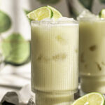 a close up shot of two a glasses of creamy coconut milk brazilian lemonade garnished with lime slices, half a cut lime at the base of the glass and ice cubes surrounding the glass.