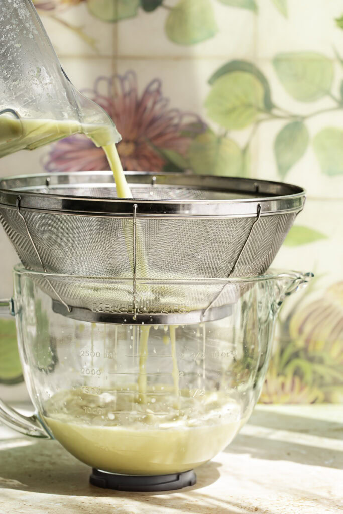 action shot straining the brazilian lemonade through a fine mesh strainer into a large glass measuring bowl.
