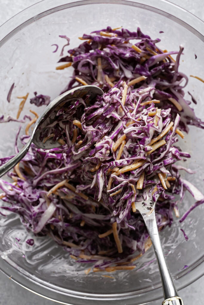 action shot of salad servers tossing the red cabbage coleslaw with the dressing.