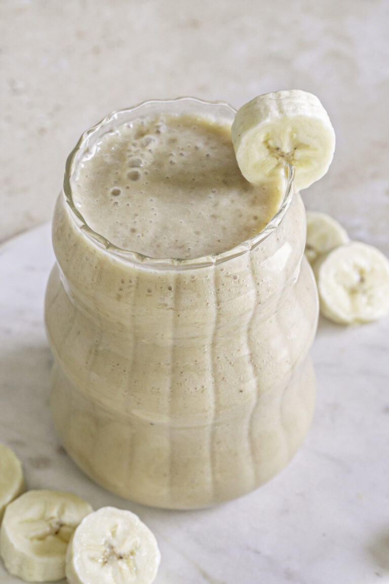 3/4 photo of banana milk in a ribbed bubble glass garnished with a slice of banana on the rim of the glass.