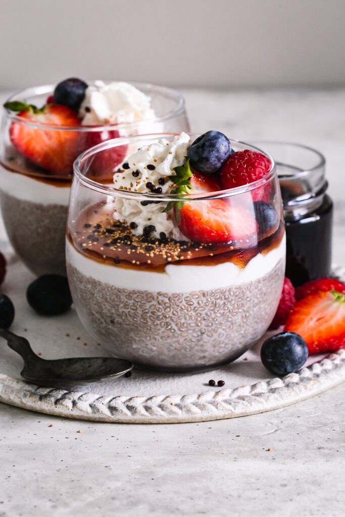 two glasses of chia seed pudding topped with coconut cream, maple syrup, whipped cream, fresh berries and chocolate covered chia seeds.