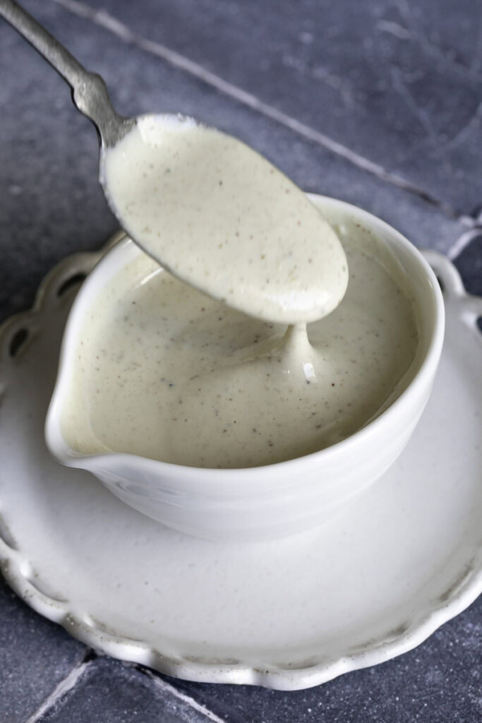 a bowl of vegan caesar dressing on a plate with a spoon drizzling some dressing back into the bowl.