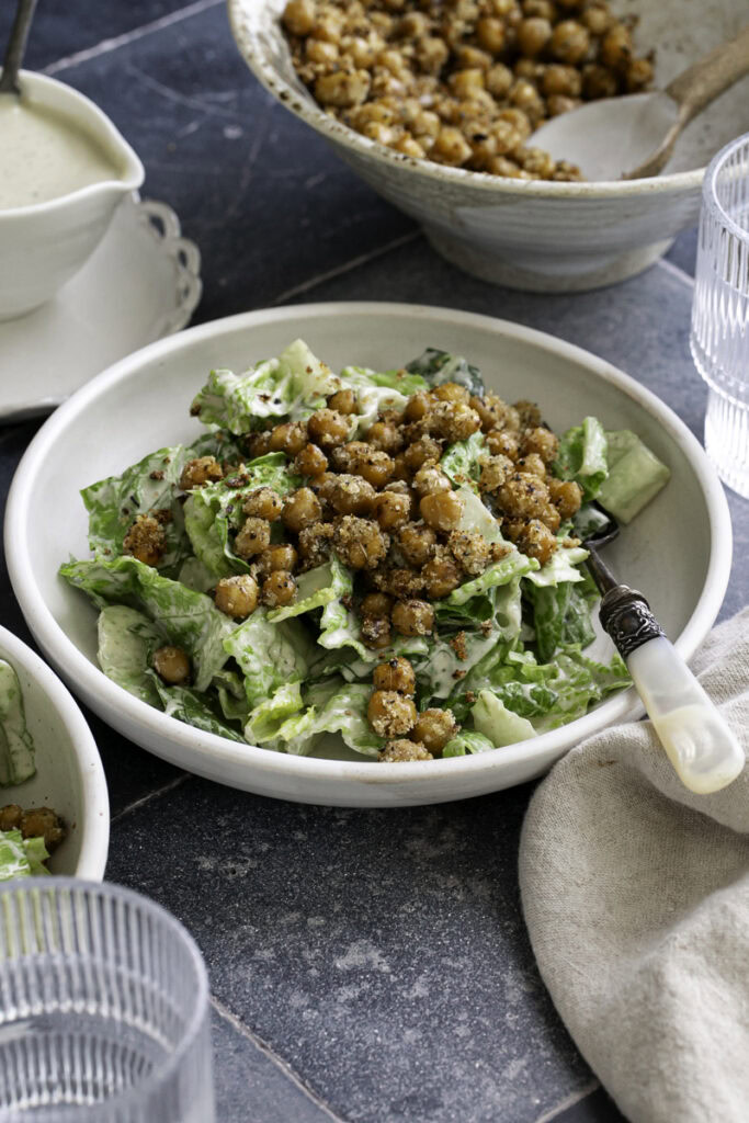 a 3/4 shot of a bowl of romaine caesar salad topped with crispy chickpea croutons, a bowl of extra caesar dressing and a bowl of extra chickpea croutons in the background.