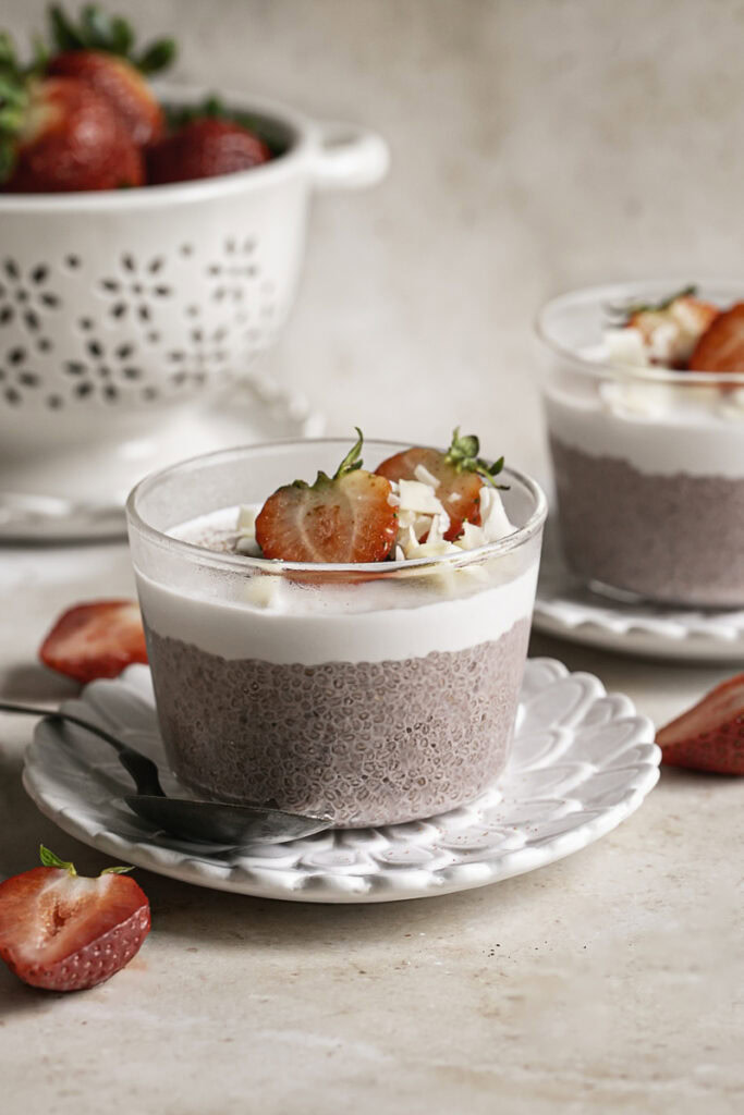 a glass filled with strawberry hibiscus chia seed pudding topped with yogurt, fresh strawberries and toasted coconut chips with another chia pudding in the background.