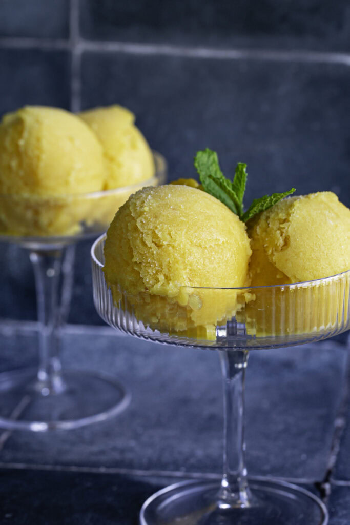 close up of two glasses filled with two scoops of mango peach frozen sorbet garnished with mint leaves.