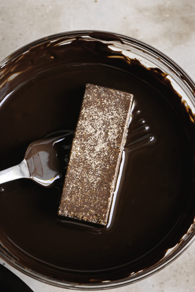a fork holding a crispy peanut butter protein chocolate bar in a bowl of melted chocolate.