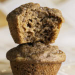 two apple cider banana bread muffins stacked on top of one another, the top muffin with a bite taken out of it.