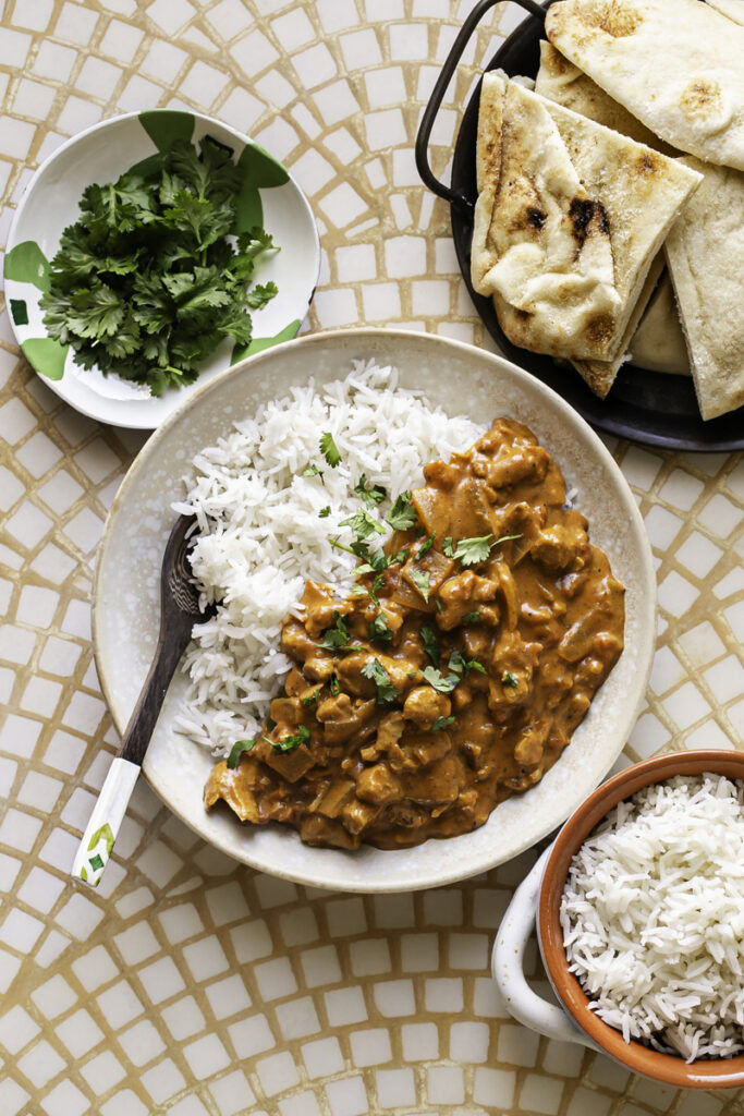 a bowl with basmati rice and indian butter chicken with a spoon in the bowl, a side bowl of rice and fresh cilantro leaves and a plate of naan.