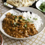 3/4 shot of a bowl with basmati rice and indian butter chicken with a spoon in the bowl, a side bowl of rice and fresh cilantro leaves and a plate of naan.