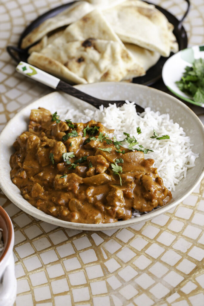 3/4 shot of a bowl with basmati rice and indian butter chicken with a spoon in the bowl, a side bowl of rice and fresh cilantro leaves and a plate of naan.