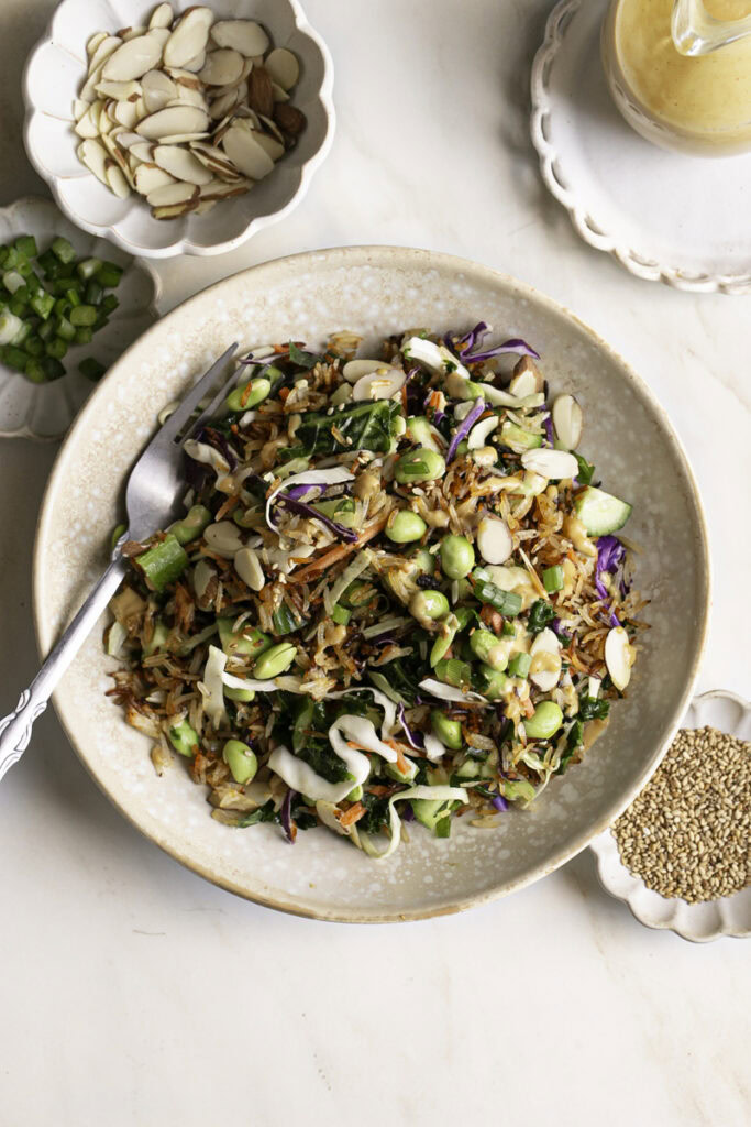 Kale Edamame Crispy Rice Salad in a bowl with a fork in it, small bowls of sliced almonds, green onions, sesame seeds and a bottle of dressing around it.