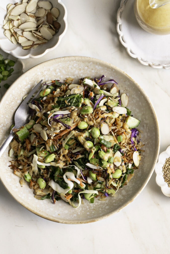 Kale Edamame Crispy Rice Salad in a bowl with a fork in it, small bowls of sliced almonds, green onions, sesame seeds and a bottle of dressing around it.