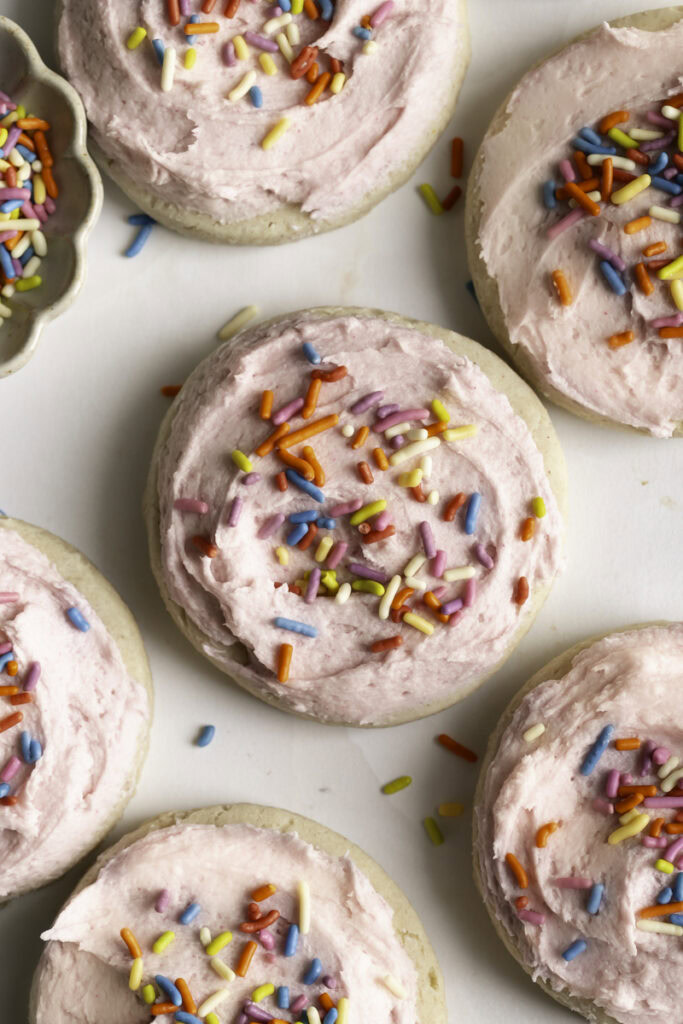 round soft frosted and sprinkled sugar cookies and a bowl of sprinkles.