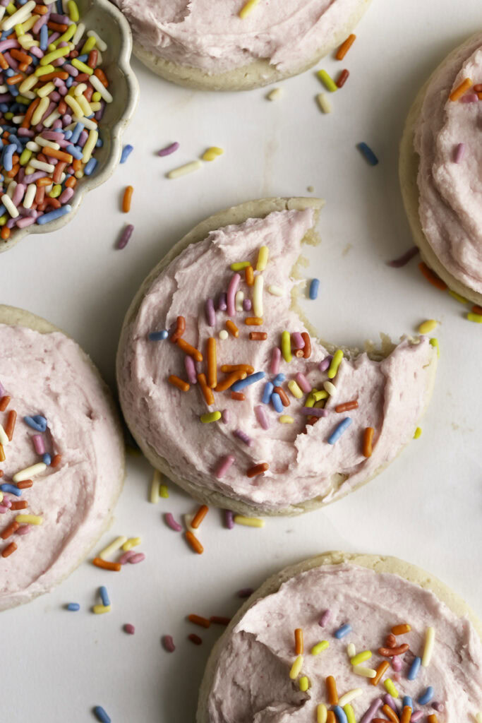 an overhead shot of soft frosted and sprinkled sugar cookies, one with a bite taken out of it and a bowl of sprinkles.