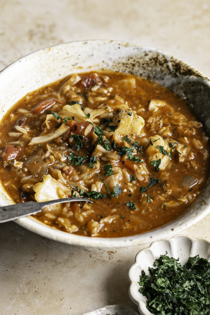 a 3/4 shot of a bowl of vegan cabbage roll soup with a spoon in the bowl and a small dish of chopped parsley.
