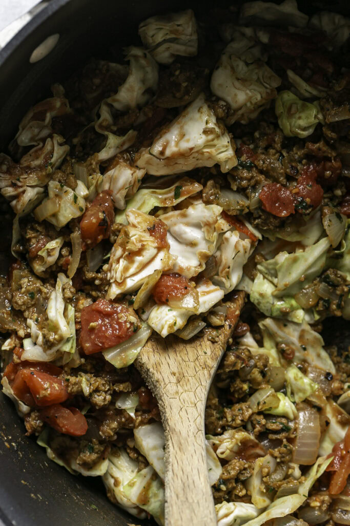a cooking pot with chopped cabbage, vegan ground beef, diced tomatoes and chopped onion with a wooden spoon.