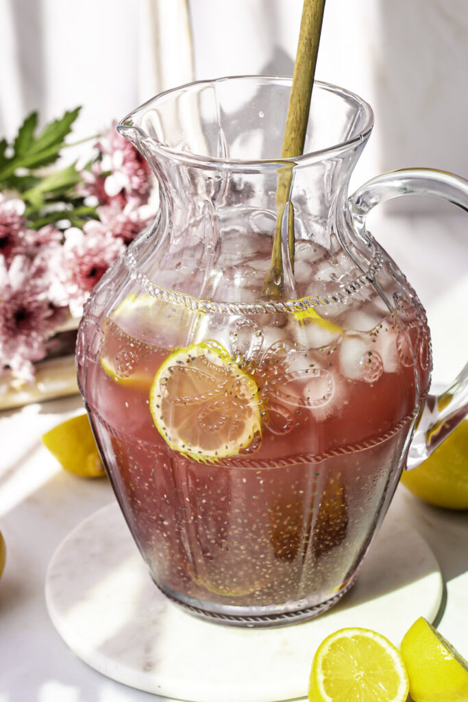 Pink Pitaya Chia Seed Lemonade in a glass pitcher with ice, lemon slices, and a wooden spoon.