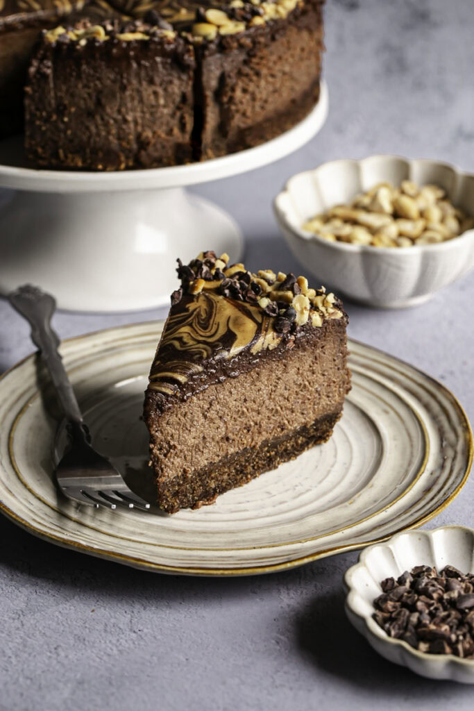 a slice of vegan triple chocolate peanut butter cheesecake on a plate with the whole cheesecake on a cake stand in the background.
