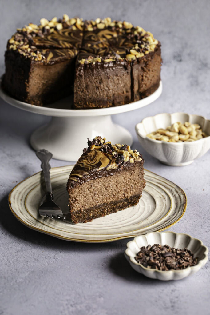 a slice of vegan triple chocolate peanut butter cheesecake on a plate with the whole cheesecake on a cake stand in the background.